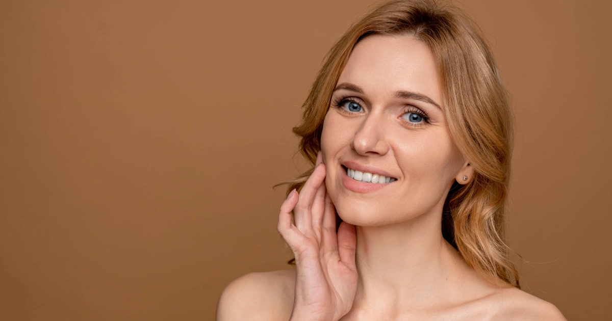 Close-up of a smiling blonde woman receiving Hydrafacial Treatment in Allen, TX, gently touching her cheek against a tan background.